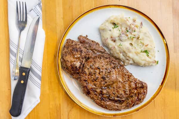 Juicy, grilled ribeye steak seasoned to perfection, plated with creamy garlic mashed red potatoes and garnished with fresh herbs on a white and gold-rimmed dish.