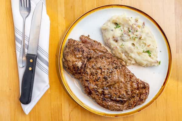 Grilled and seasoned ribeye steak served with creamy garlic mashed potatoes, garnished with herbs on a white ceramic plate.
