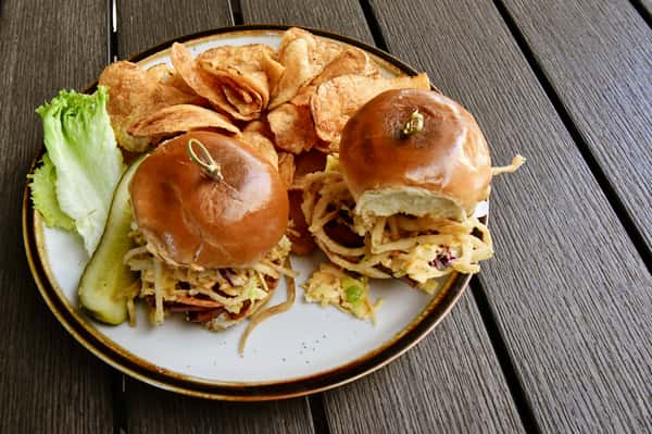 Two slider sandwiches with crispy onion straws and slaw on soft brioche buns, served with house-made potato chips and a pickle spear on a rustic plate.
