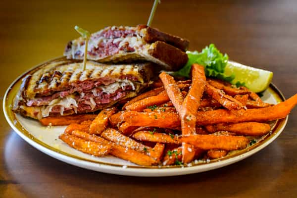 Grilled Reuben sandwich stacked with corned beef, sauerkraut, and melted cheese on rye, served with seasoned sweet potato fries and a pickle wedge.