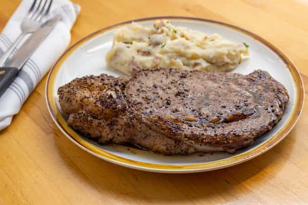 Juicy peppercorn-seasoned ribeye steak served with creamy mashed red potatoes, garnished with fresh herbs on a white plate with gold trim.