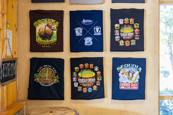 Display of colorful Sequoia Brewing Company graphic t-shirts mounted on a wall, showcasing various beer logos and local brew artwork inside a rustic taproom.
