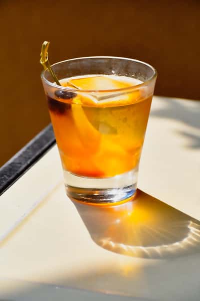 Craft Old Fashioned cocktail served in a lowball glass with a large ice cube, garnished with an orange peel and cherry, casting a warm shadow in natural light.