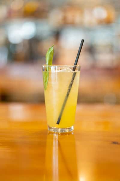 Refreshing yellow cocktail served in a clear glass with a black straw and fresh basil garnish, set against a warm wooden bar background.