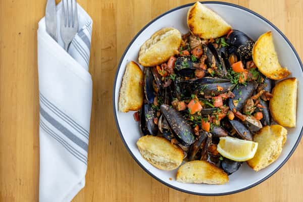 Bowl of steamed black mussels topped with diced tomatoes, fresh herbs, and crumbled bacon, served with toasted garlic bread and a lemon wedge on a rustic wooden table.