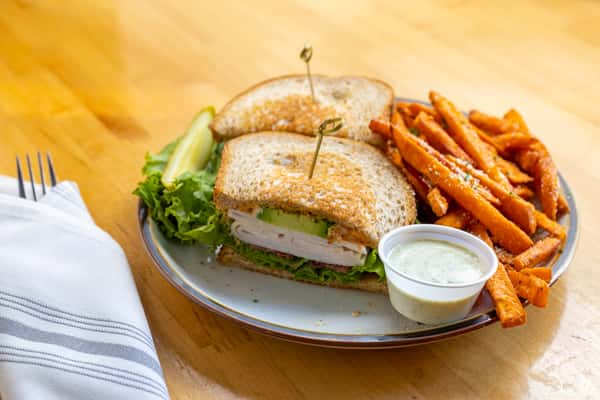 Toasted turkey and avocado sandwich layered with lettuce on whole grain bread, served with crispy sweet potato fries, dill pickle spear, and creamy ranch dip.