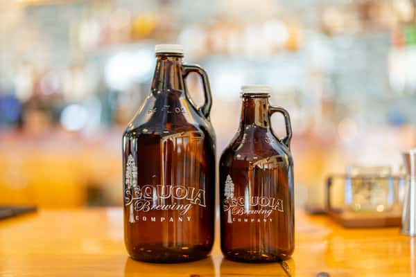 Large and small brown glass growlers featuring the Sequoia Brewing Company logo, displayed on a polished bar counter.