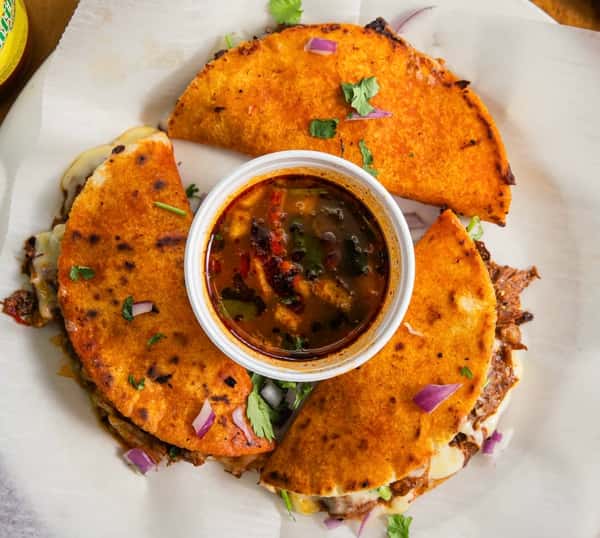 Pinches Tacos: Three orange birria tacos with melted cheese and a cup of dipping consommé on white paper.