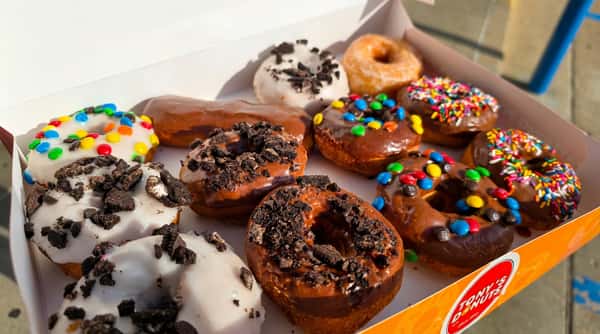 Assorted donuts in a white box, including chocolate, white icing with sprinkles and cookie crumbles, and a plain glazed donut.