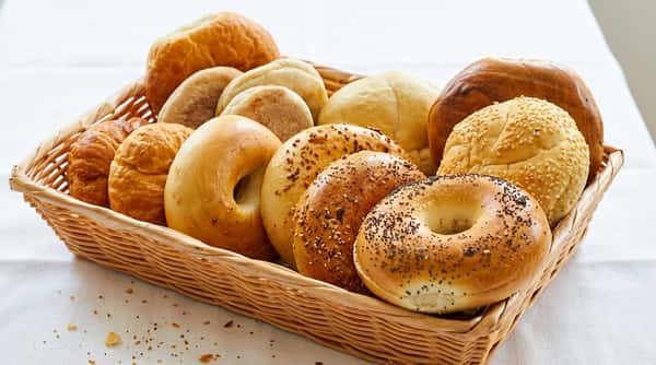 A basket filled with assorted baked goods, including bagels with poppy seeds and sesame seeds, plain bagels, croissants, English muffins, and bread rolls.