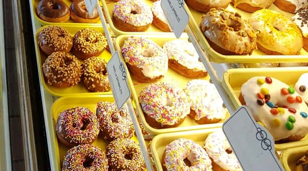 Assorted donuts in a display case; some with chocolate and nuts, vanilla with multi-colored sprinkles, and white icing with M&M's.