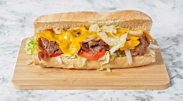 Cheesesteak sandwich on a hoagie roll with steak, melted cheddar, lettuce, onions, and tomato on a cutting board.