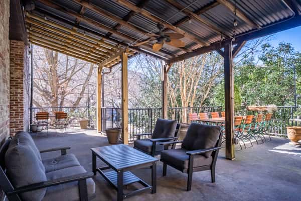 Covered outdoor patio featuring cushioned lounge seating and wooden dining tables surrounded by trees and natural light. A relaxing, shaded space perfect for enjoying casual food and craft beers in a laid-back setting.