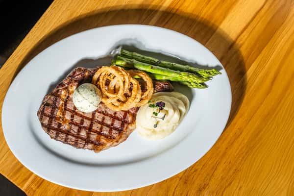 Overhead view of a grilled ribeye steak topped with herb butter and crispy onion rings, served with creamy mashed potatoes and charred asparagus. A steakhouse favorite featuring bold, classic flavors.