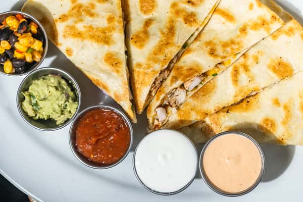 Close-up of grilled chicken quesadilla slices served with guacamole, salsa, sour cream, chipotle mayo, and corn-black bean salsa. A flavorful Tex-Mex appetizer perfect for sharing.
