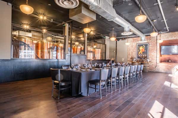 Elegant private dining room at Max Lager's with a long banquet table set with dinnerware, wine glasses, and black linens. Large copper brewing tanks are visible through glass panels, blending industrial brewery charm with upscale event space ambiance.