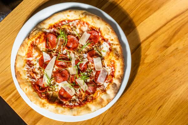 Top-down view of a classic pepperoni pizza with tomato sauce, mozzarella, basil, and parmesan shavings on a bubbly wood-fired crust. A crowd-pleasing favorite at any pizzeria or American grill.