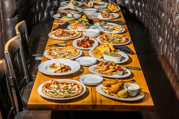 A long wooden table filled with Max Lager's signature dishes, including wood-fired pizzas, burgers, fried shrimp, spinach artichoke dip, grilled steaks, and more. Set in a tufted leather booth dining space, this feast showcases the full range of American grill and brewery favorites.