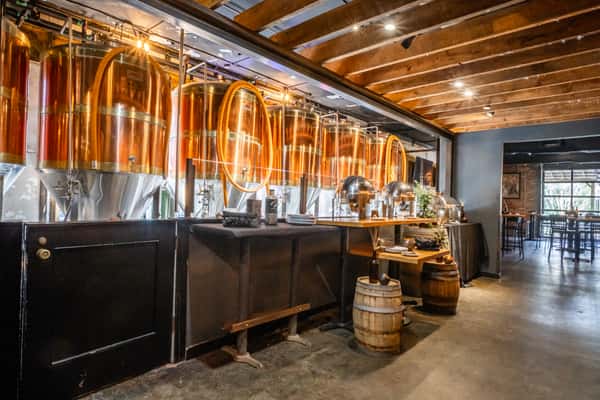 Row of gleaming copper brewing tanks visible behind a buffet station set with chafing dishes and rustic barrels. The brewery ambiance blends industrial design with warm, welcoming hospitality, ideal for events or private dining.