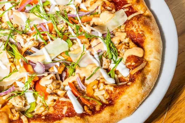 Close-up of a wood-fired pizza topped with grilled chicken, red onions, bell peppers, shaved parmesan, and drizzles of white and orange sauces, garnished with basil. A spicy and savory pizza bursting with flavor.