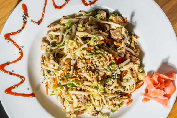 Asian-style chicken noodle salad with shredded vegetables, grilled chicken, and sesame seeds, drizzled with sweet chili sauce and served with pickled ginger on a white plate. A bold, flavorful twist on salad classics.