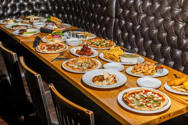 Long wooden table filled with an array of dishes including pizzas, ribs, shrimp, salads, and more, set against tufted leather seating. A rich display of American grill and brewpub fare in a warm dining environment.