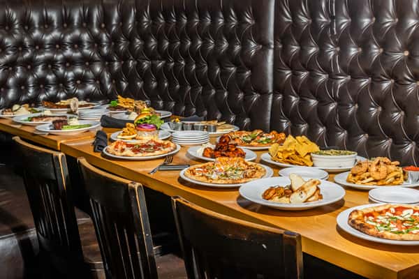 Long booth table lined with signature dishes including wood-fired pizzas, burgers, fried shrimp, wings, and quesadillas at Max Lager's. A hearty showcase of American grill favorites in a cozy tufted leather setting.