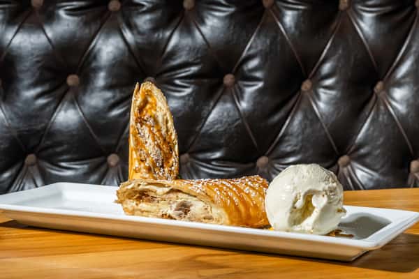 Warm apple strudel filled with spiced apples and flaky pastry, drizzled with caramel sauce and lightly dusted with powdered sugar, served with a scoop of creamy vanilla ice cream on a rectangular white plate. A comforting and indulgent dessert.