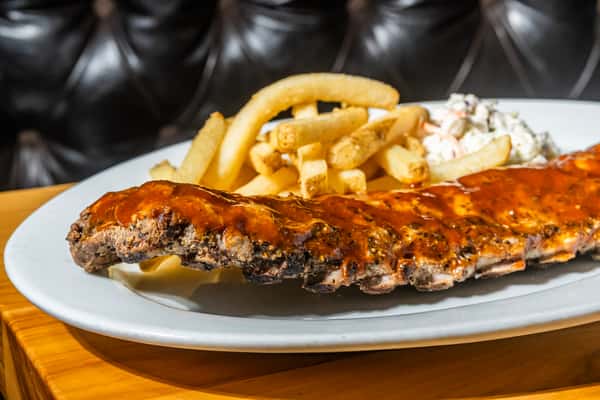 Full rack of barbecue ribs glazed in rich sauce, served with seasoned French fries and creamy coleslaw on a white oval plate. A hearty BBQ platter ideal for casual dining at an American grill.