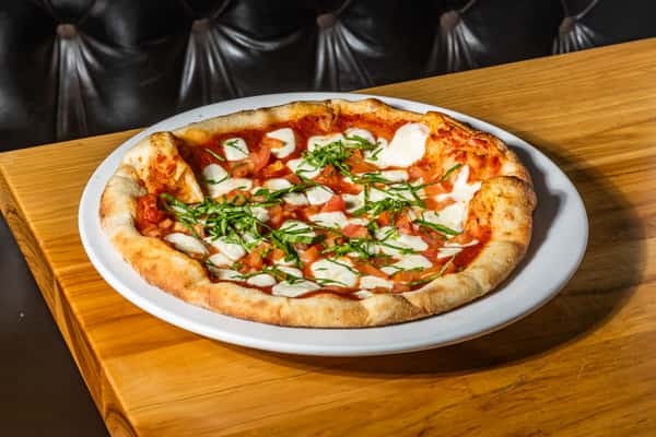 Wood-fired Margherita pizza topped with fresh mozzarella, diced tomatoes, and thinly sliced basil over rich tomato sauce on a golden, blistered crust. A classic Italian-American favorite served on a white plate.