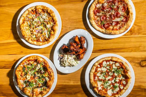 Overhead view of assorted personal-size pizzas and barbecue wings with coleslaw on a wooden table. Toppings include pepperoni, fresh mozzarella, basil, and vegetables for a flavor-packed dining experience.