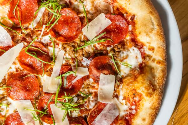 Close-up of a wood-fired pepperoni pizza topped with shaved parmesan, fresh basil, and melted mozzarella on a crispy golden crust. A savory slice of Italian-American comfort food.