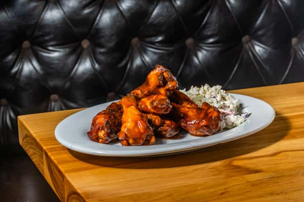 Stacked barbecue chicken wings coated in smoky sauce, paired with creamy coleslaw on a white plate. A saucy, satisfying appetizer in a cozy booth setting.