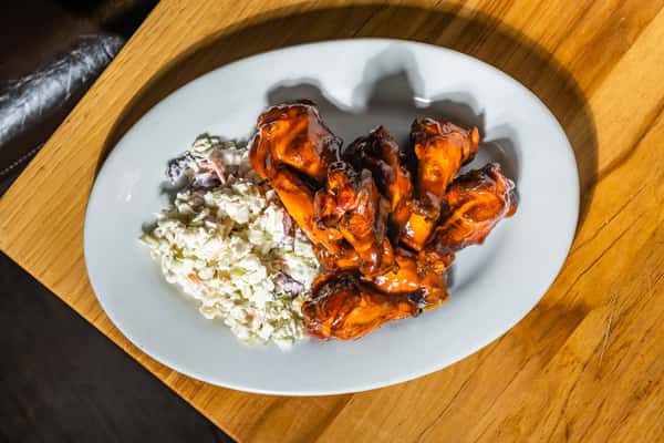 Overhead view of saucy barbecue wings and creamy coleslaw on a white oval plate, served on a wooden table. A bold appetizer highlighting smoky and creamy contrasts.