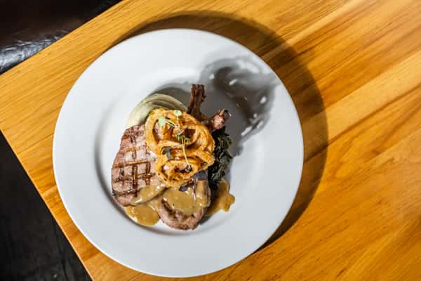 Overhead shot of a grilled pork chop topped with fried onion rings and gravy, served with mashed potatoes and braised greens. A savory, Southern-inspired plate on a rustic wooden table.