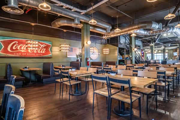Spacious industrial-style dining room featuring wood tables, metal chairs, and green walls adorned with vintage Coca-Cola and American flag signage. Exposed ductwork and pendant lighting complete the casual, nostalgic brewpub ambiance.