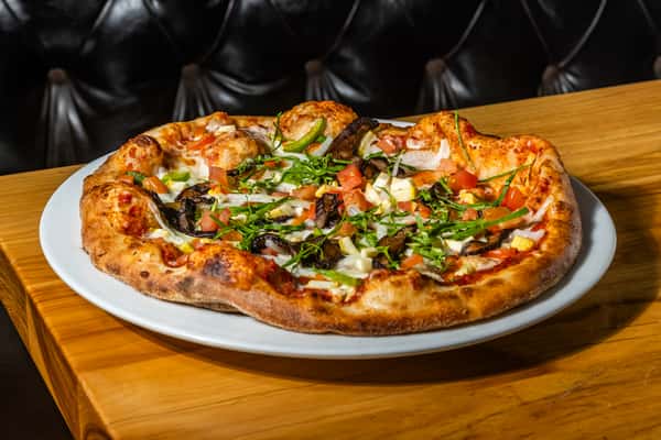 Wood-fired veggie pizza topped with mushrooms, diced tomatoes, onions, mozzarella, and fresh basil on a golden, blistered crust. A garden-fresh option for pizza lovers.