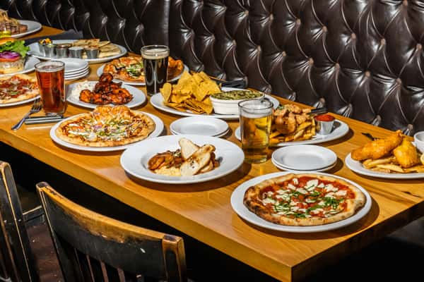 Generous spread of Max Lager's signature dishes including pizzas, fried fish, grilled meats, dips, and more on a wooden table in a tufted leather booth setting. A feast showcasing diverse American grill and brewery favorites.
