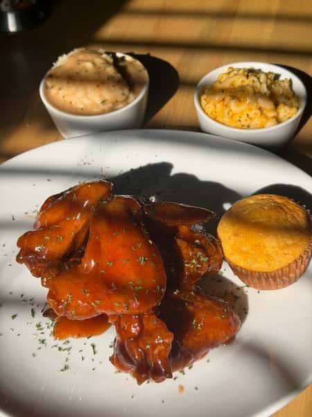 Glazed BBQ chicken served with creamy mashed potatoes, baked macaroni and cheese, and a golden cornbread muffin on a white plate.