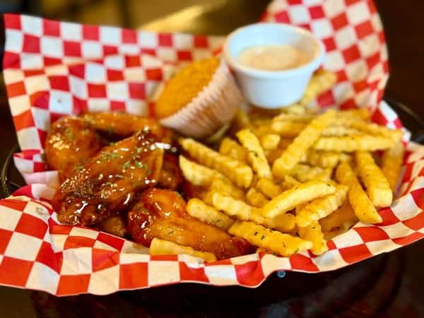 Crispy glazed chicken wings served in a red checkered basket with crinkle-cut fries, cornbread muffin, and dipping sauce for a classic comfort food combo.