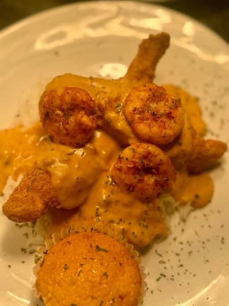 Southern fried catfish topped with Cajun cream shrimp sauce, served with a cornbread muffin on a white plate.