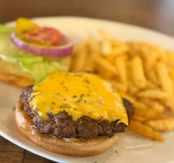 Classic cheeseburger with melted cheddar on a toasted bun, served open-faced with crisp lettuce, tomato, red onion, and golden French fries on a white plate.
