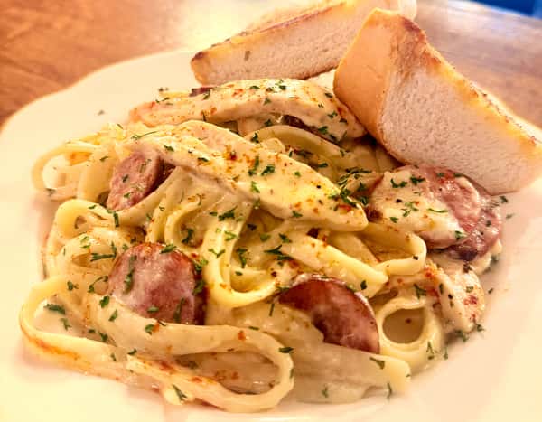 Creamy Cajun chicken and smoked sausage fettuccine Alfredo, tossed with herbs and topped with grilled chicken slices, served with toasted garlic bread on a white plate.