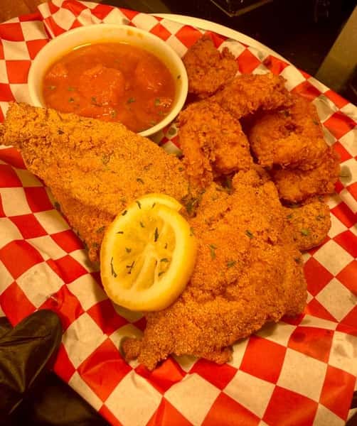 Southern fried catfish platter with crispy breaded fillets, golden hush puppies, lemon slices, and a side of tangy dipping sauce in a red checkered basket at a seafood restaurant.