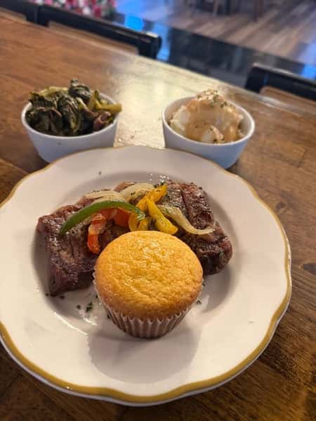 Grilled steak topped with sautéed bell peppers and onions, served with a golden cornbread muffin, collard greens, and creamy mashed potatoes in a casual Southern restaurant setting.