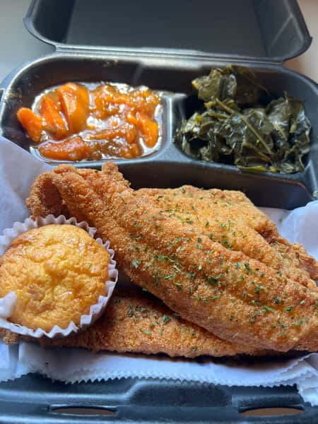 Southern fried catfish fillets with cornbread muffin, collard greens, and candied yams in a takeout container, showcasing classic soul food comfort cuisine.