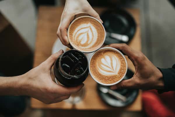 Three people toast with two cups of latte art and one glass of iced coffee over a wooden café table with plates and cutlery.