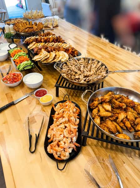 Catering buffet spread with shrimp platter, chicken wings, grilled skewers, tacos, fresh vegetables, and dipping sauces arranged on a wooden table for party catering.