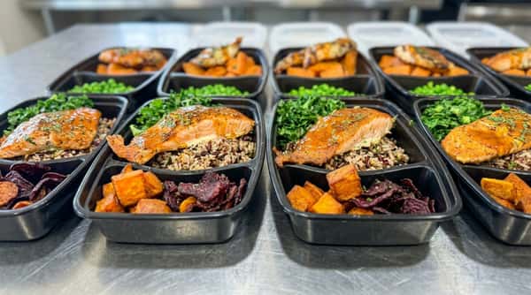 Healthy salmon meal prep containers with wild rice, roasted sweet potatoes, kale, peas, and roasted vegetables arranged in black takeout containers for healthy meal delivery or catering.