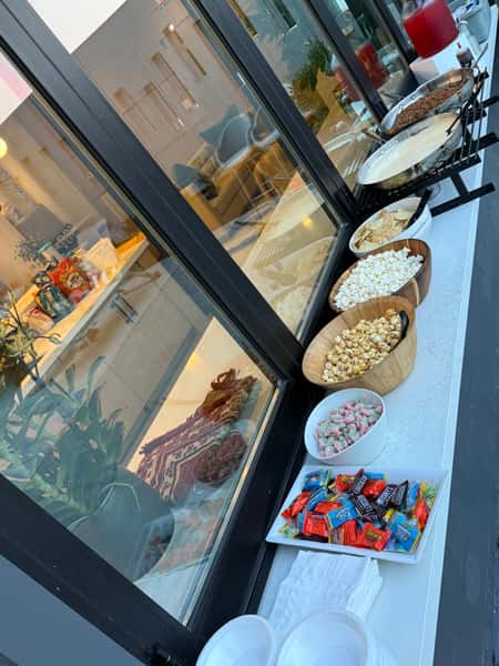 Event snack station with bowls of popcorn, chips, candy, and sweet treats arranged on a buffet table near large windows, casual self-serve dessert and snack bar for a party or reception.